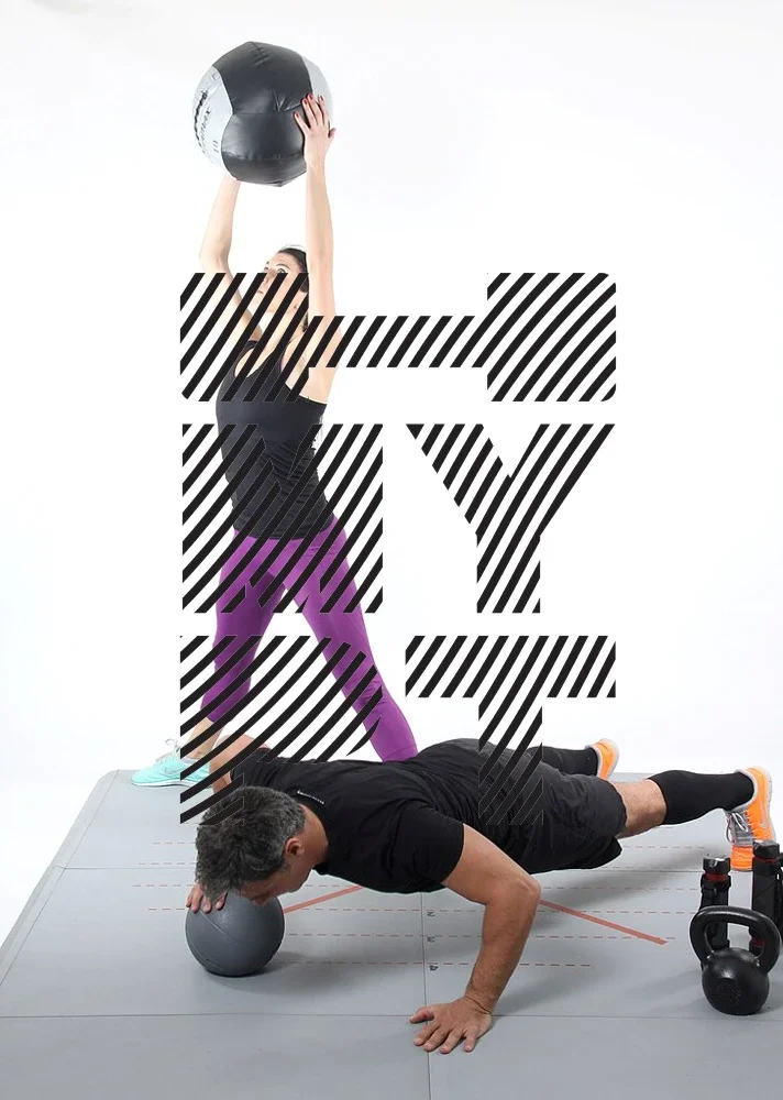 Two people performing functional fitness exercises; a woman standing upright, holding a medicine ball overhead, and a man performing a push-up with one hand elevated on a smaller medicine ball. Fitness equipment like a kettlebell is visible nearby.