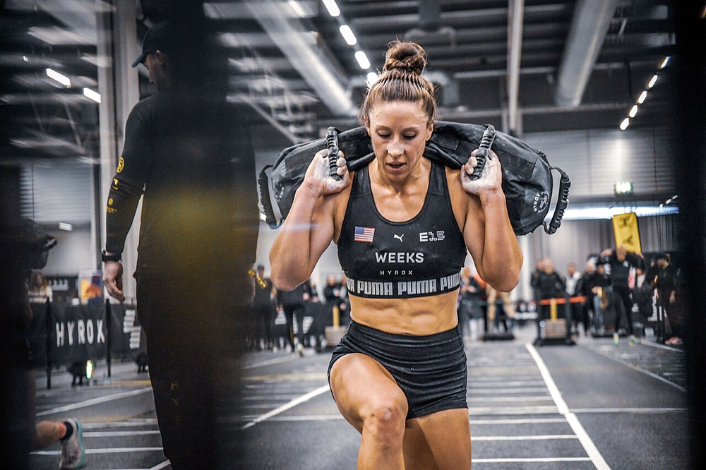 Female athlete performing weighted walking lunges with a sandbag during a HYROX competition, showcasing glute strength and endurance.