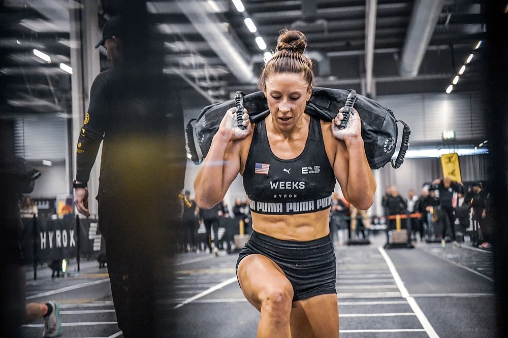 Female athlete performing weighted walking lunges with a sandbag during a HYROX competition, showcasing glute strength and endurance.